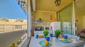 Un balcón con una mesa de comedor lista para dos personas, un refrigerador, un microondas y una pequeña área de cocina con cafetera y objetos decorativos.