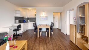 Una cocina moderna con armarios blancos y un microondas, un comedor con una mesa y sillas de madera, y un mueble de madera con puertas de cristal a la derecha.