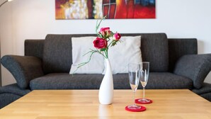 Un salón moderno con un sofá gris, un jarrón blanco con flores rosas y dos vasos sobre una mesa de madera.