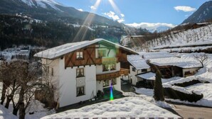 Ein traditionelles Chalet mit Balkon, umgeben von schneebedeckten Bergen und blauem Himmel.