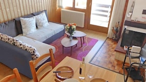 Una sala de estar acogedora con un sofá, una mesa de comedor con sillas, una mesita redonda con un jarrón de flores y una chimenea.
