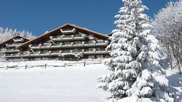 Un edificio de varios pisos con fachada de madera y balcones, rodeado de árboles cubiertos de nieve y un cielo azul despejado.