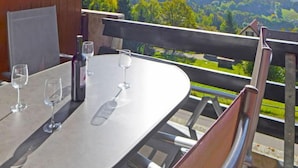 Una mesa con cuatro copas de vino y una botella, en un balcón con vistas a un valle boscoso.