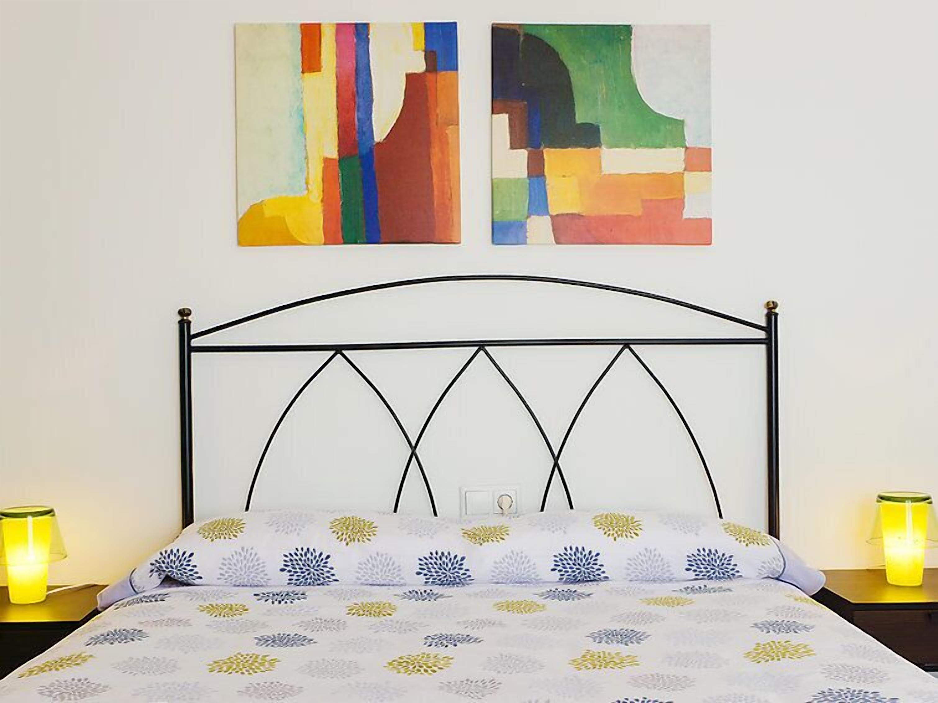 Een bed met een wit beddengoed met bloemenprint, een zwart metalen hoofdbord en twee bedlampen met gele lampenkappen.