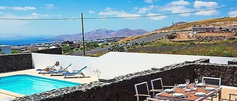 Una piscina con sillas de descanso y un comedor con mesa y sillas, con vistas a un paisaje costero con montañas al fondo.