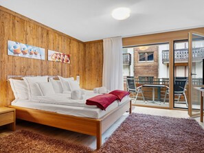 A modern bedroom with a wooden headboard, a large bed with white linens, a red throw blanket, and a view of a balcony with outdoor furniture. - Rütschi by Interhome (Zermatt)