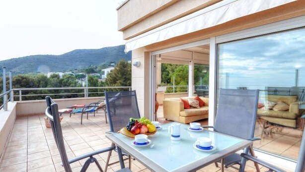 A balcony with a table set for two, chairs, a sofa, and a glass door leading to an outdoor seating area. - Apartment Fane by Interhome (Llançà)