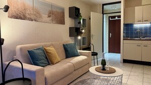 A modern living room with a beige sofa, a round coffee table, and a kitchen area with blue tiles and a sink.