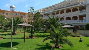 Une cour d’hôtel agrémentée de palmiers, d’un parasol en chaume et d’un bâtiment doté de nombreux balcons.