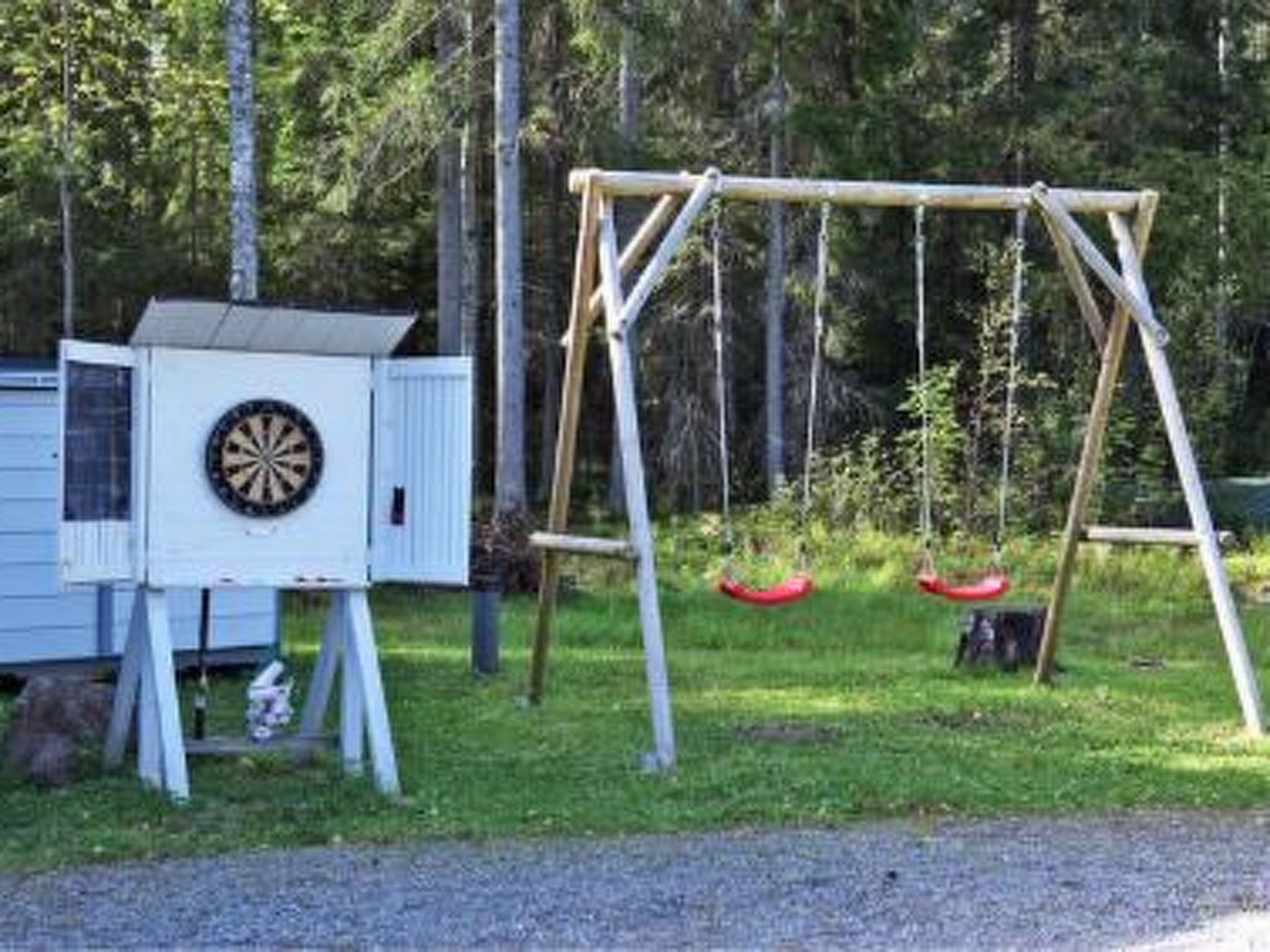 Puisesta materiaalista valmistettu keinuteline, jossa on kaksi keinu, pieni varasto, jonka sisällä on tikanheitotaulu, sekä aidattu alue, jonka taustalla on puita.