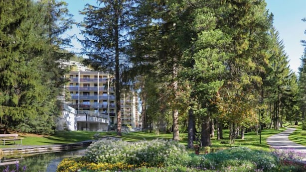 A landscaped garden with a pond, surrounded by dense trees and a multi-story building in the background. - Allod Park Haus C 106 by Interhome (Davos)