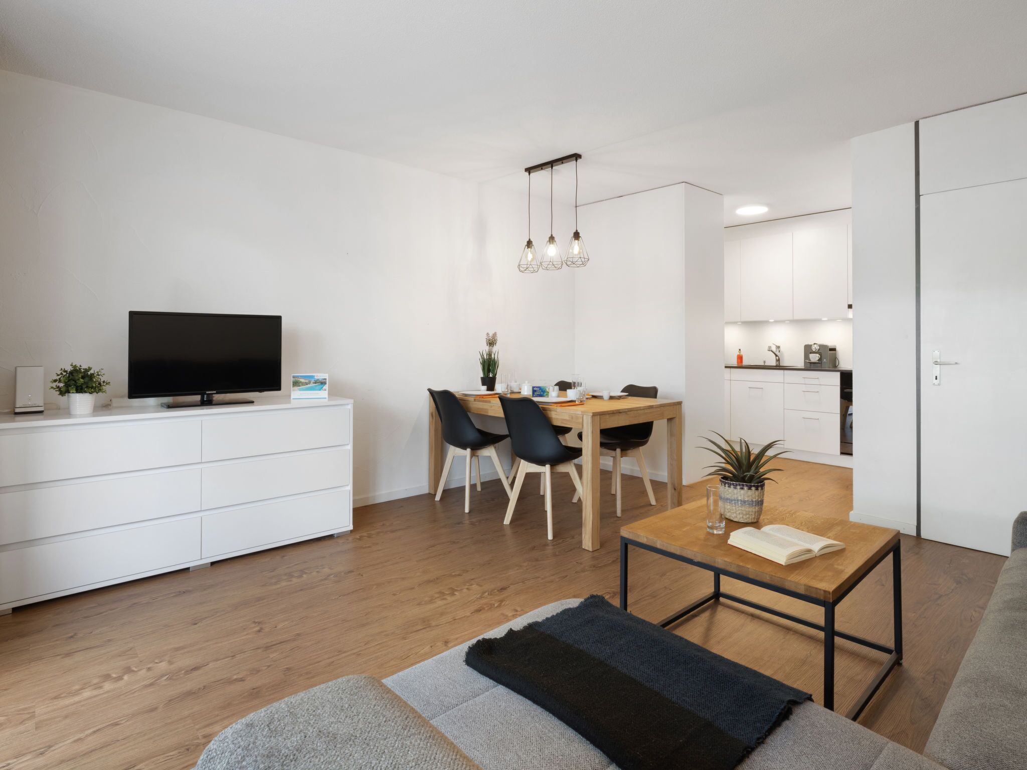 Una sala de estar moderna y minimalista con un televisor de pantalla plana, un comedor con mesa y sillas de madera, y una cocina con armarios blancos.