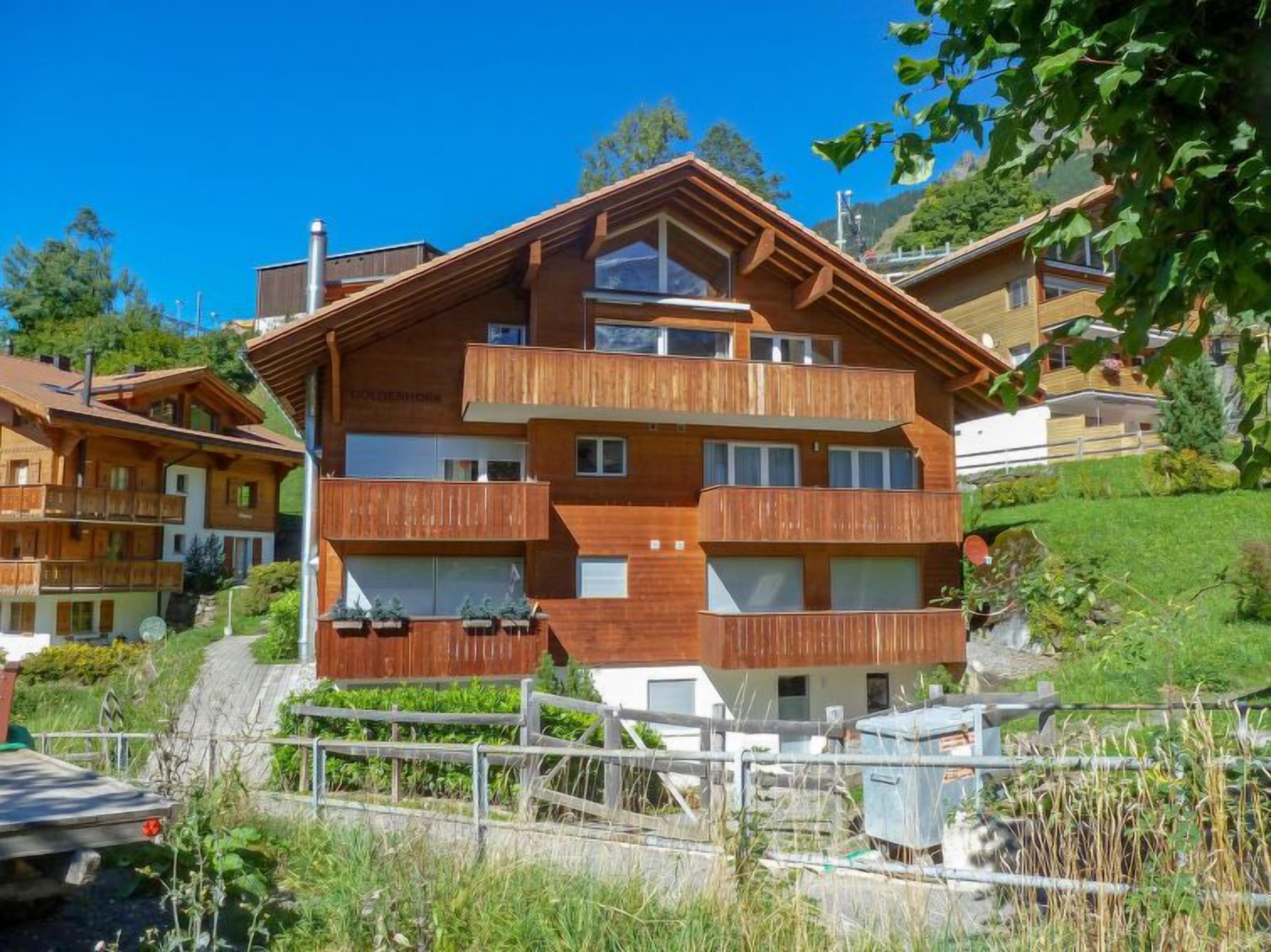 En flervånings chalet i trä med balkonger, omgiven av frodig grönska och andra byggnader i chaletstil.