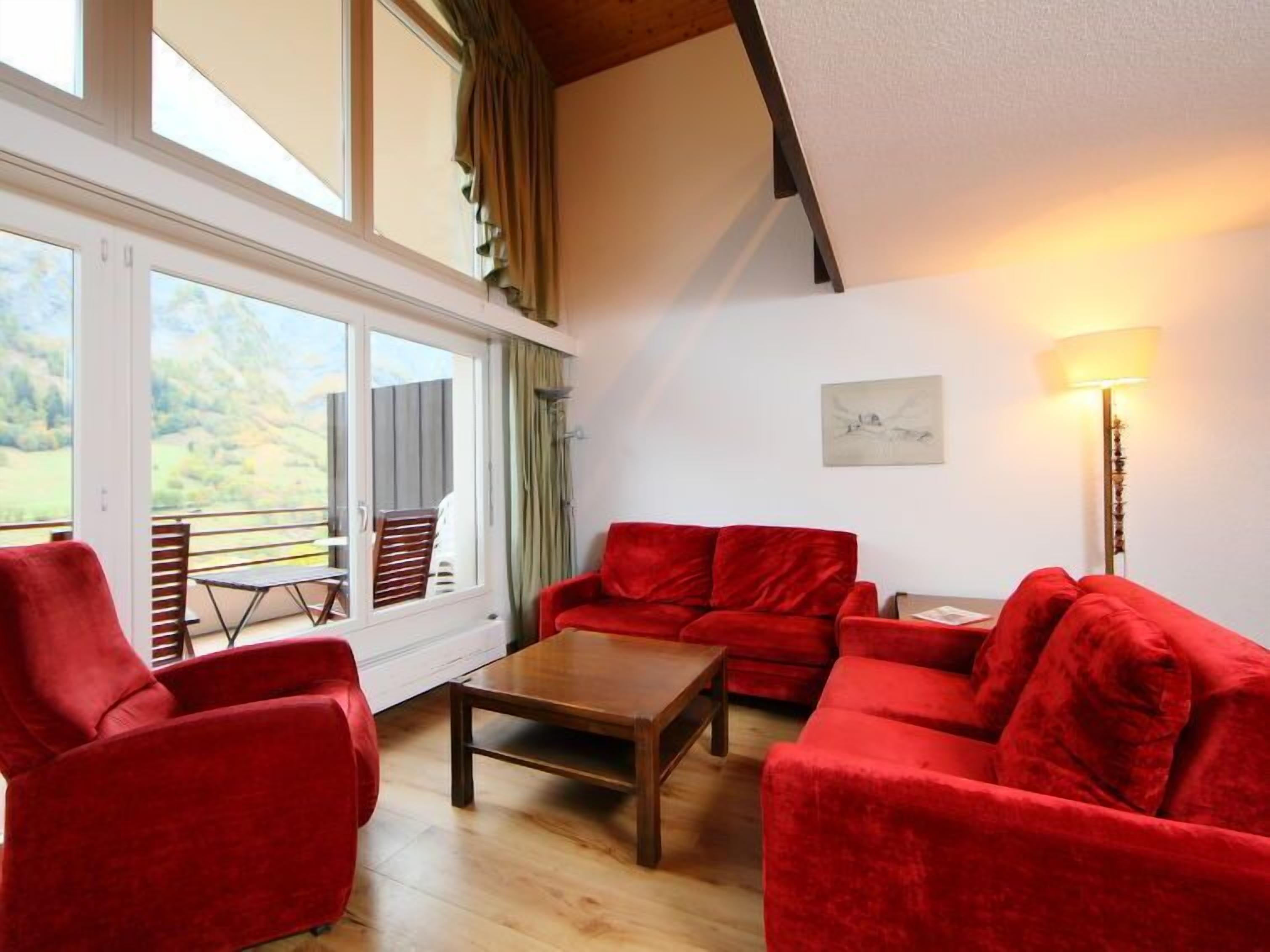 Ein geräumiges Wohnzimmer mit einer roten Sofagarnitur, einem Holztisch und einem Esstisch mit Stühlen. Große Fenster bieten einen Blick ins Freie.