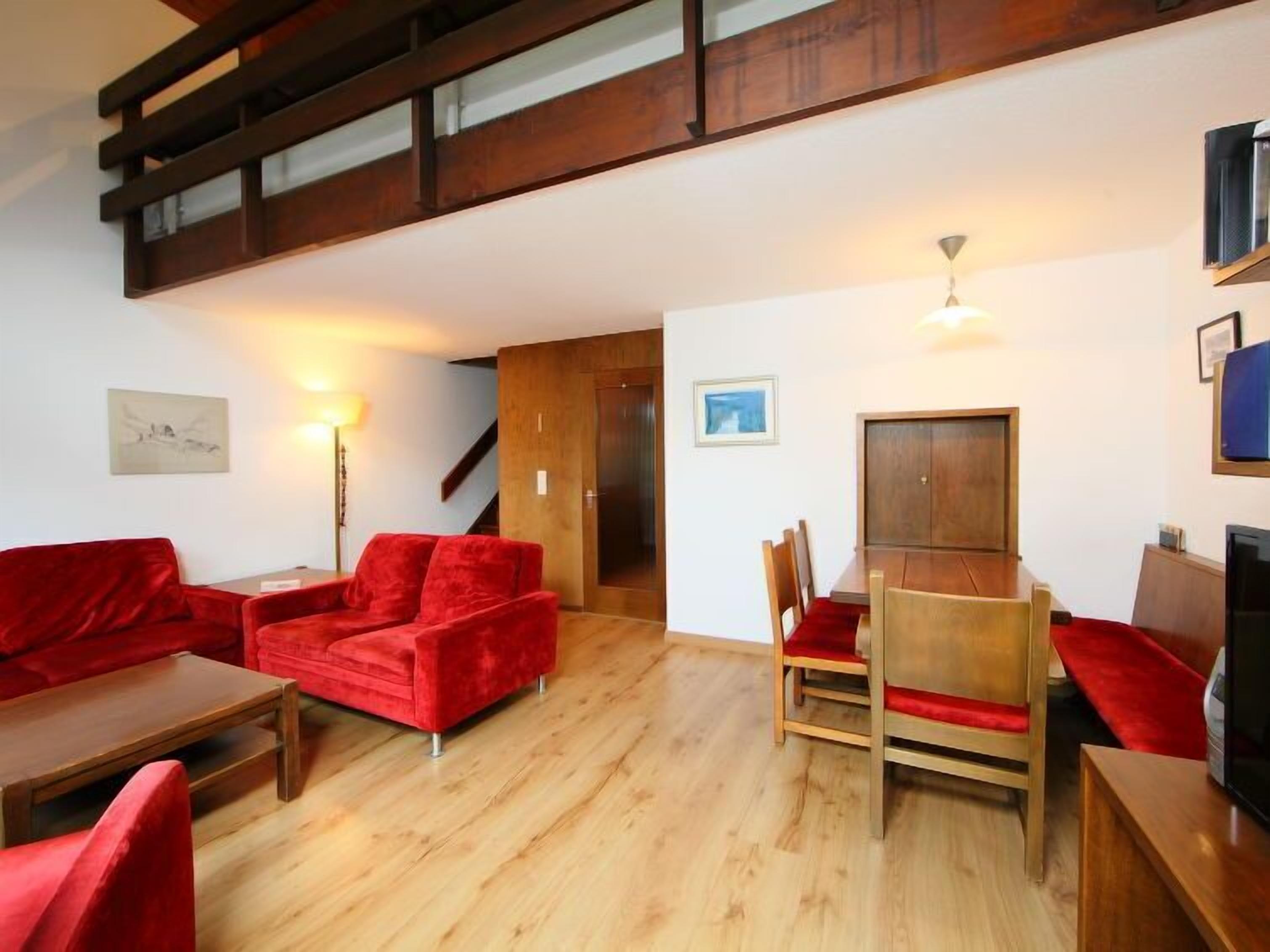 A spacious living room with a red sofa, wooden dining table, and chairs. A staircase leads to an upper level.