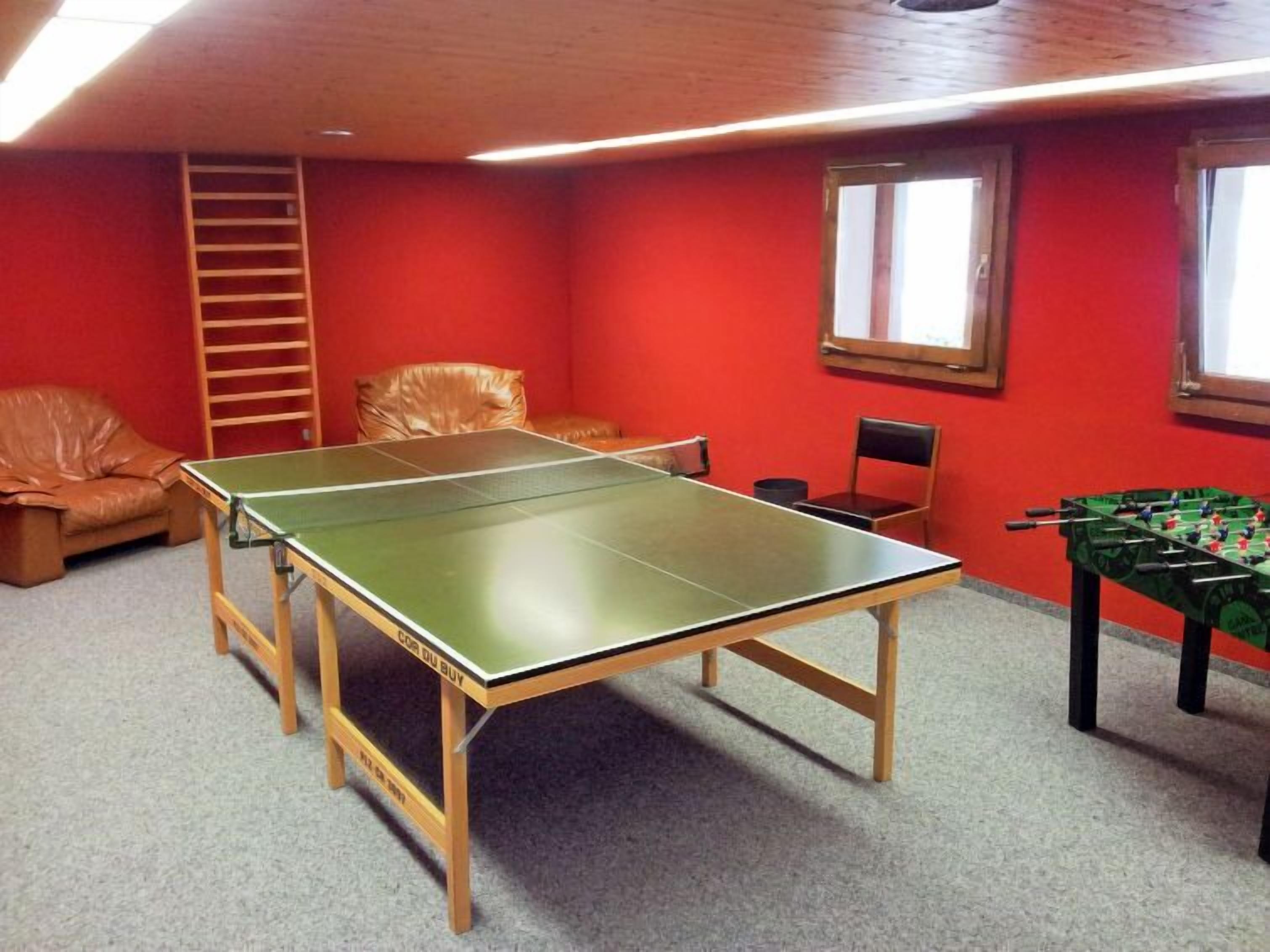 A room with a ping pong table, a foosball table, a brown leather chair, and a wooden ladder against a red wall.