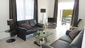 A modern living room with a black leather sofa, a glass coffee table, a flat-screen TV, and a dining area with a table and chairs. - Plevier Comfort by Interhome (Noordwijk)