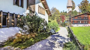 A traditional building with a steeply pitched roof, a visible clock tower, and a paved pathway leading to a grassy area. - Primula Apt. B by Interhome (Wengen)