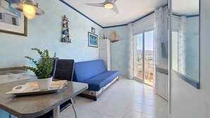 Een kamer met een blauwe bank, een eettafel met een plant, een plafondventilator en uitzicht door een glazen deur.