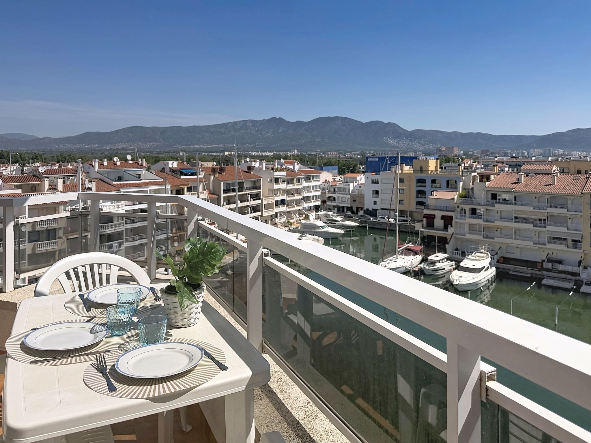 Un balcón con una mesa lista para comer, con vistas a un puerto deportivo con barcos y un paisaje urbano con montañas al fondo.