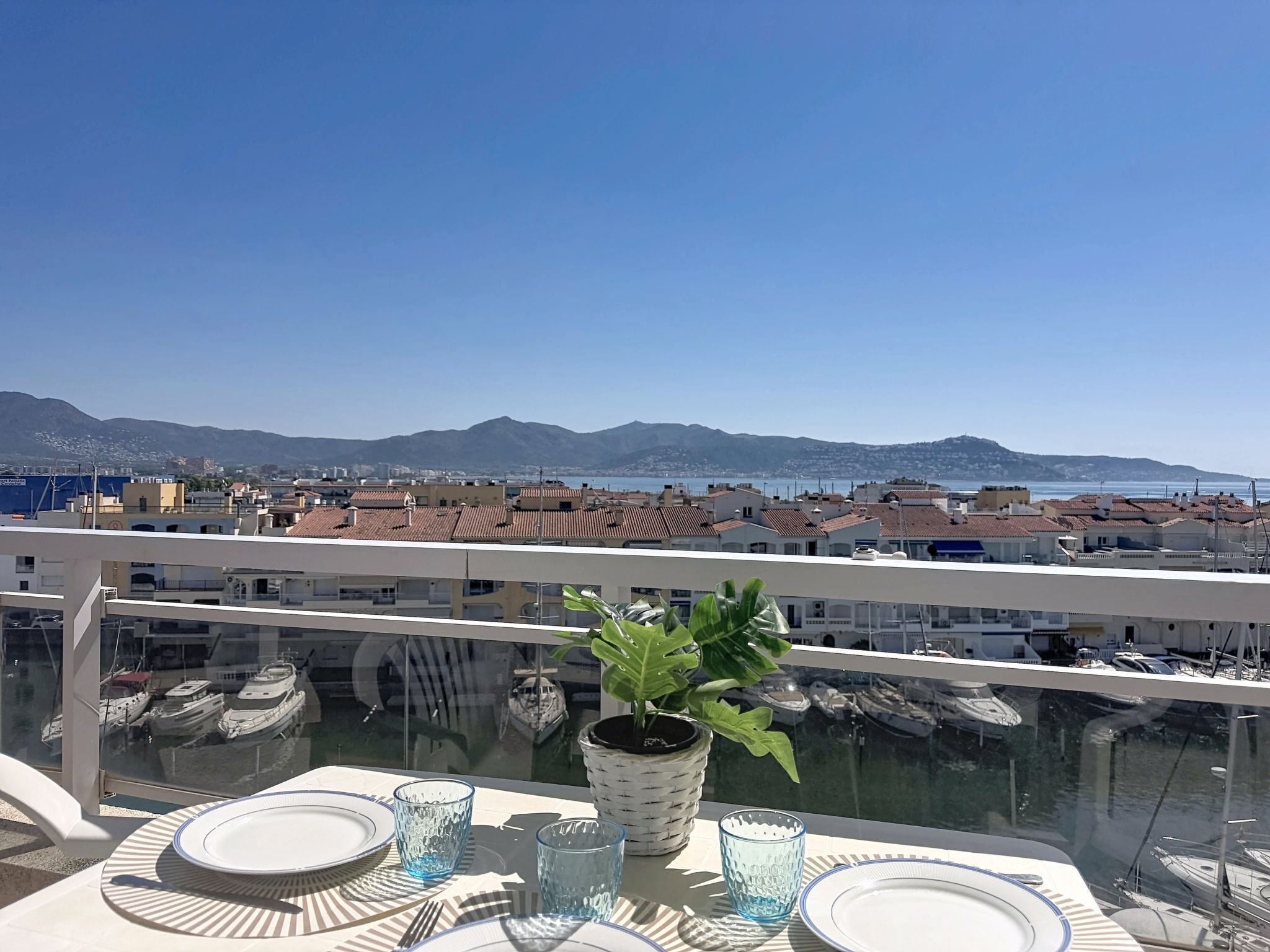 Un balcon avec une table dressée pour le dîner, donnant sur une marina où sont amarrés des bateaux, et sur une chaîne de montagnes lointaine.