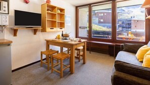 Ein kleines Hotelzimmer mit einem Holztisch, zwei Stühlen, einem Fernseher, einem Fenster mit Blick auf Gebäude und einem Sofa mit gelben Kissen.