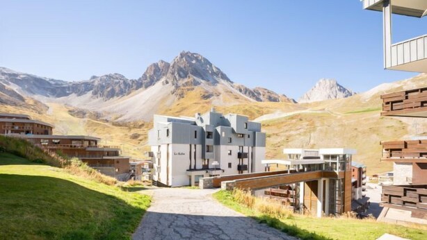 Ein modernes Gebäude mit einem zentralen Eingang, umgeben von einem gepflasterten Weg und Grünflächen, vor einer Bergkulisse.