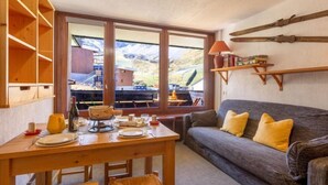 Ein gemütlicher Essbereich mit einem Holztisch, gedeckt für zwei Personen, einer Couch mit gelben Kissen und Blick auf eine gebirgige Landschaft durch große Fenster.