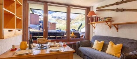 A cozy dining area with a wooden table set for two, a sofa with yellow cushions, and a view of a mountainous landscape through large windows.