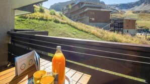Eine Terrasse mit einer hölzernen Bank, einem Tisch mit Orangensaft und einer Speisekarte. Die Terrasse bietet Blick auf eine gebirgige Landschaft mit Gebäuden im Hintergrund.