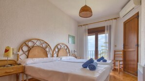 Een slaapkamer met twee bedden, een houten hoofdeinde, een nachtkastje met een lamp en uitzicht op zee door een raam.