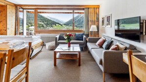 Une chambre d’hôtel moderne dotée d’une grande fenêtre donnant sur les montagnes, d’un téléviseur à écran plat, d’un canapé, d’une table basse et d’un lit en bois.