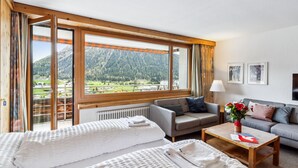 Une chambre d’hôtel dotée d’une grande fenêtre donnant sur les montagnes, d’un lit avec une literie blanche, d’un canapé gris et d’une petite table sur laquelle est posé un vase de fleurs.