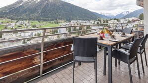 Un balcon avec une table et des chaises en bois, d’où l’on peut admirer un paysage montagneux et une ville située en contrebas.