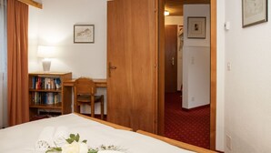 Una habitación de hotel con una cama, un escritorio, una silla, una estantería y una puerta que da a otra habitación.