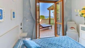 Un dormitorio con cama, mesita de noche, lámpara, vista al mar y balcón con una silla.