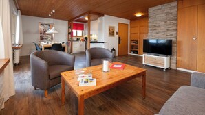 A living room with a wooden coffee table, two armchairs, a sofa, a TV, and a kitchen in the background. - Chalet Bienli Mitte by Interhome (Grindelwald)