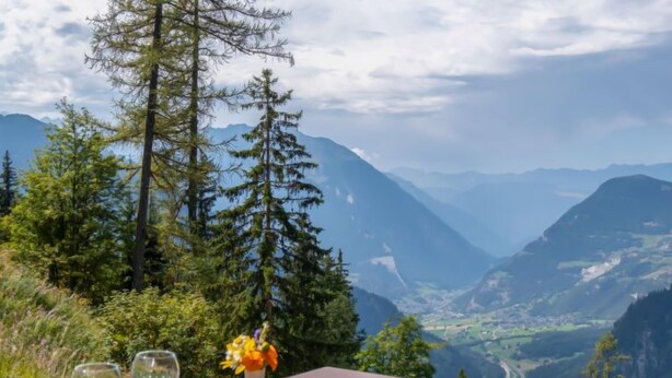A wooden deck with two empty wine glasses and a vase of flowers, overlooking a mountainous landscape.