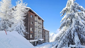 A multi-story building with balconies, surrounded by snow-covered trees and a clear blue sky.