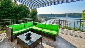 A covered outdoor seating area with a green sofa and a glass coffee table, overlooking a lake with boats. - Apartment Marija by Interhome (Krk/Omišalj)