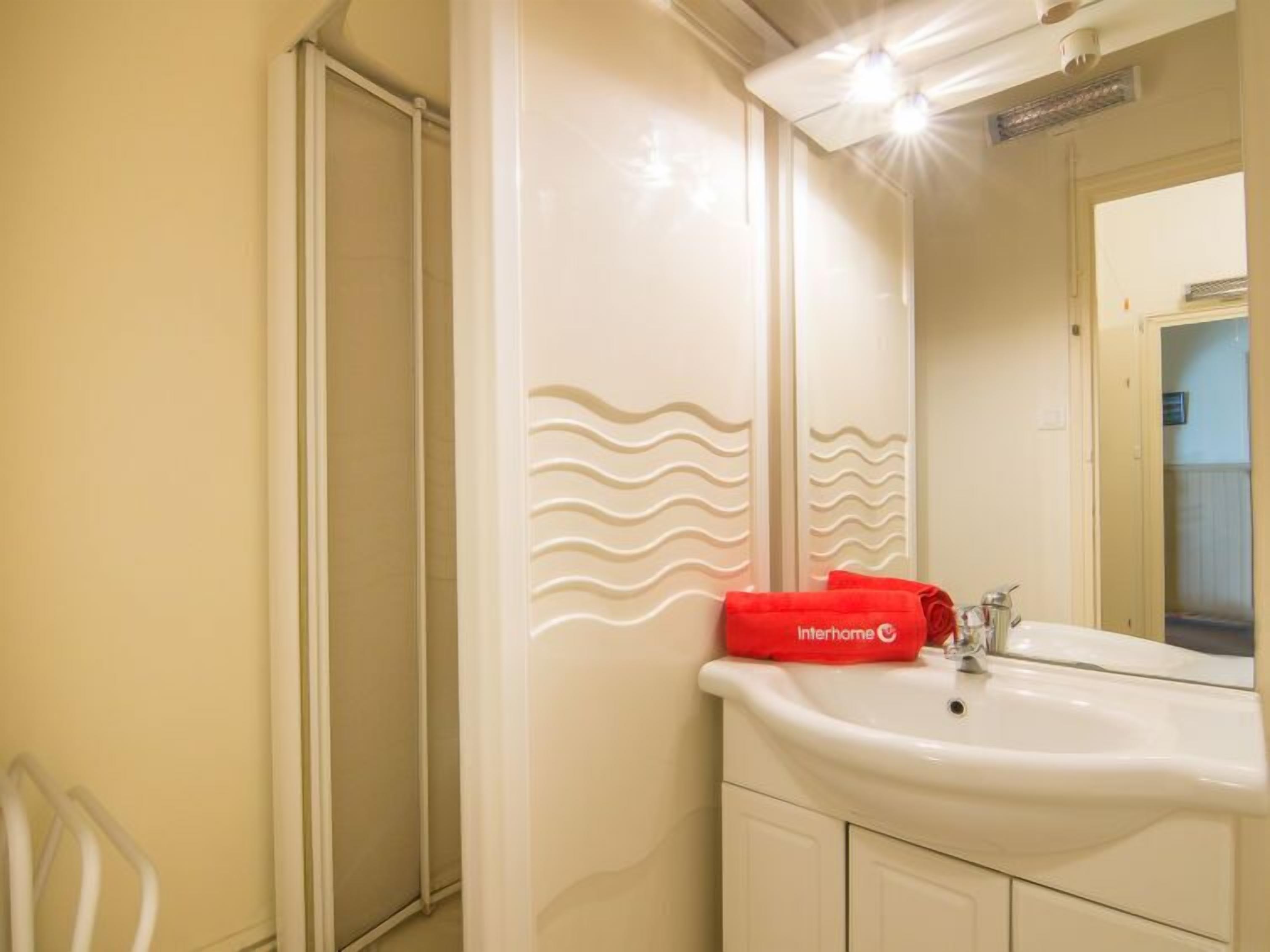 Une salle de bain avec un lavabo blanc, un miroir et une douche. Une serviette rouge de la marque Interhome est posée sur le lavabo.