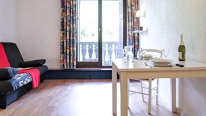 Ein Zimmer mit einer schwarzen Couch, einem Holzesstisch und einem Balkon mit gemusterten Vorhängen.