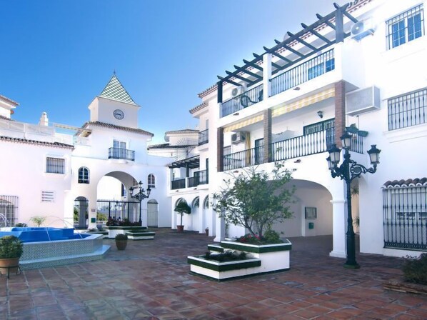 A courtyard with white buildings, a clock tower, and a blue swimming pool.