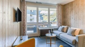 A room with a sofa, a small round table, a TV mounted on the wall, and large windows with curtains. - Le Pramecou (Val Claret) by Interhome (Tignes)