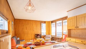 Ein gemütliches Zimmer mit Holzwänden, einem Esstisch mit Orangen und einem Buch, einem Fernseher und einem Fenster mit Vorhängen.