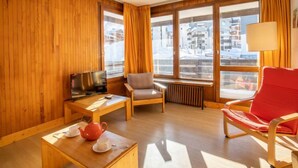 Ein gemütliches Zimmer mit Holzwänden, einem Fernseher, einem roten Sessel, einem Holztisch mit Teekanne und Tasse sowie großen Fenstern mit Vorhängen.