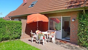 Una casa con tetto in tegole rosse, facciata in mattoni e un patio con sedie bianche e un ombrellone arancione.