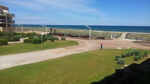 Een kustgezicht met een verhard pad dat naar een strand leidt, omringd door groen en een modern gebouw.