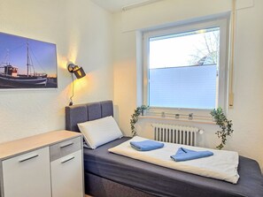 Un dormitorio con cama, escritorio, radiador, ventana con persianas y una lámpara de pared, además de un cuadro enmarcado.