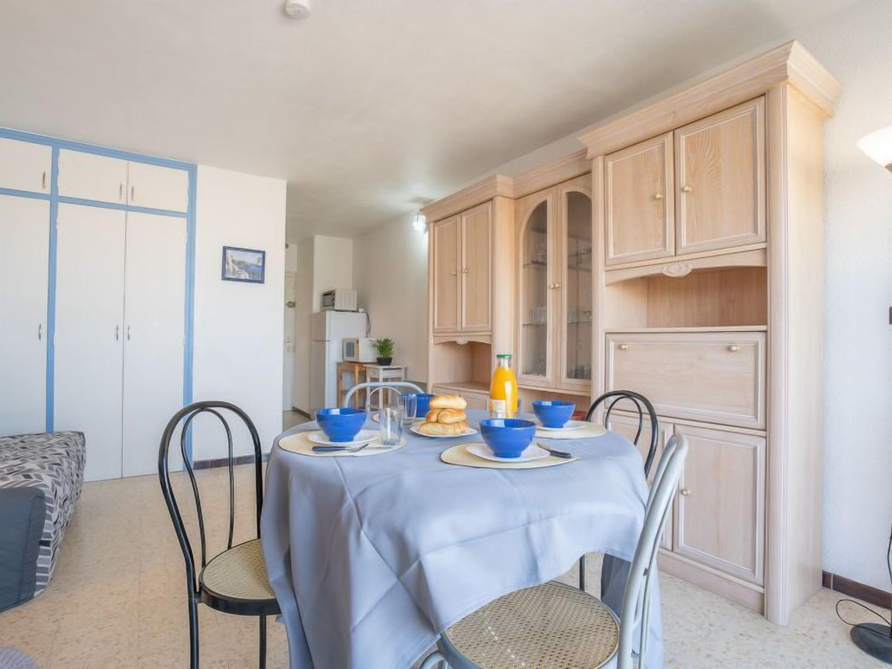 A dining area with a table set for four, surrounded by chairs. There are cabinets with glass doors and a small kitchenette in the background.